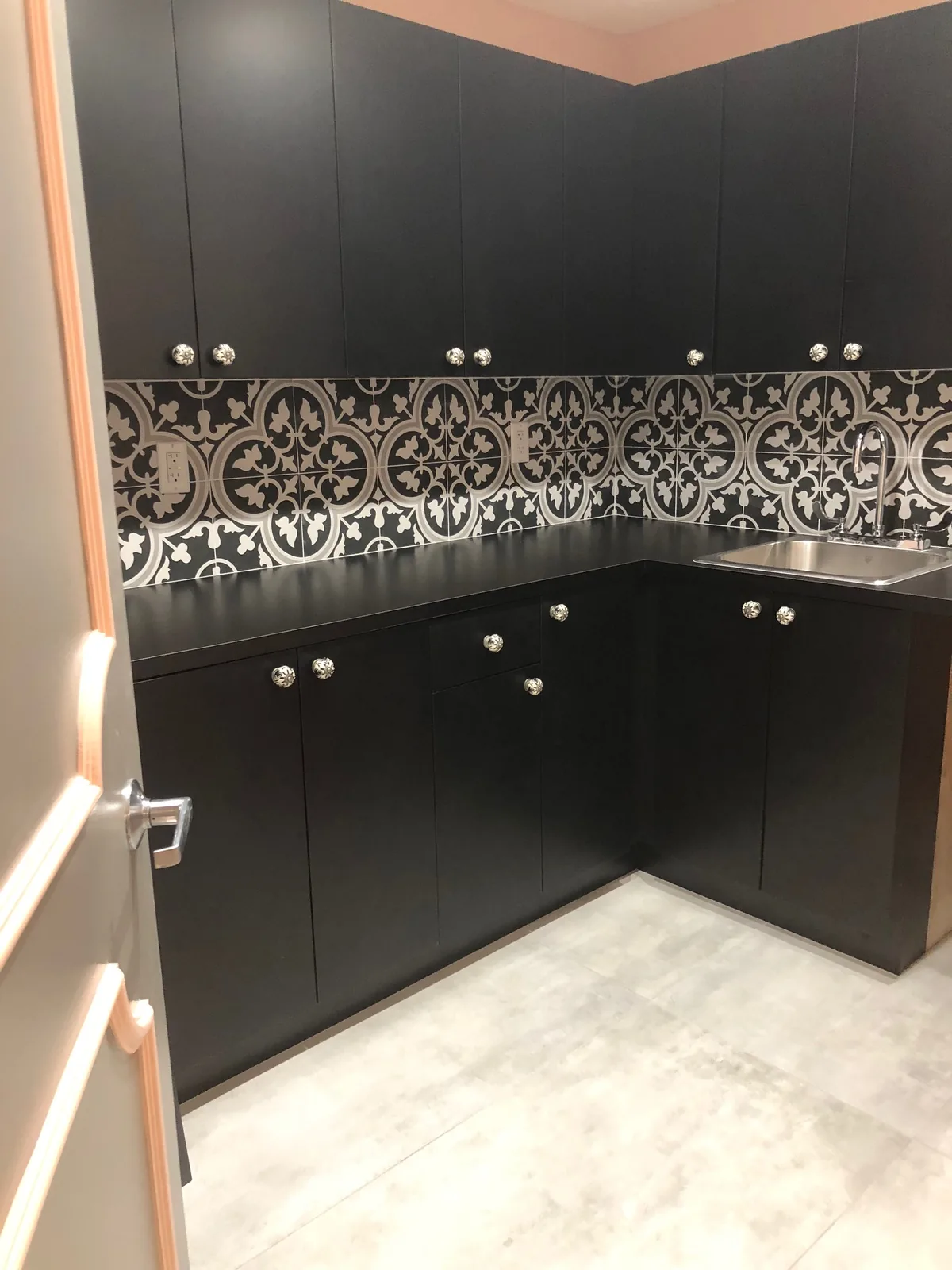 Black kitchen cabinetry with ornate patterned backsplash and stainless countertop.