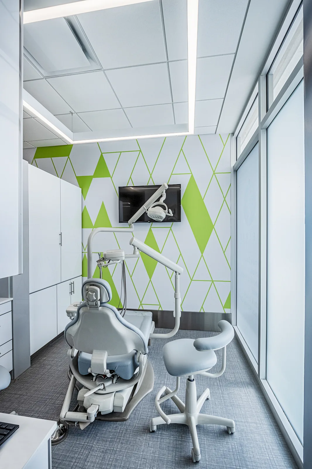 Dental operatory with white equipment, geometric green pattern, overhead monitor.